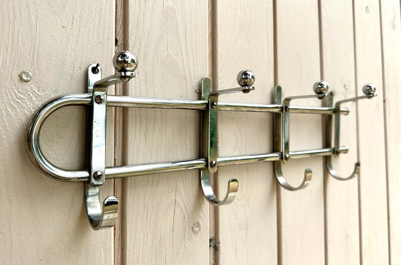 Puede incluir: Un perchero de pared fabricado en metal con acabado cromado pulido. El soporte presenta una estructura horizontal de doble barra con cuatro ganchos orientados hacia abajo y cuatro perchas orientadas hacia arriba, cada una rematada con un peque&ntilde;o remate esf&eacute;rico.