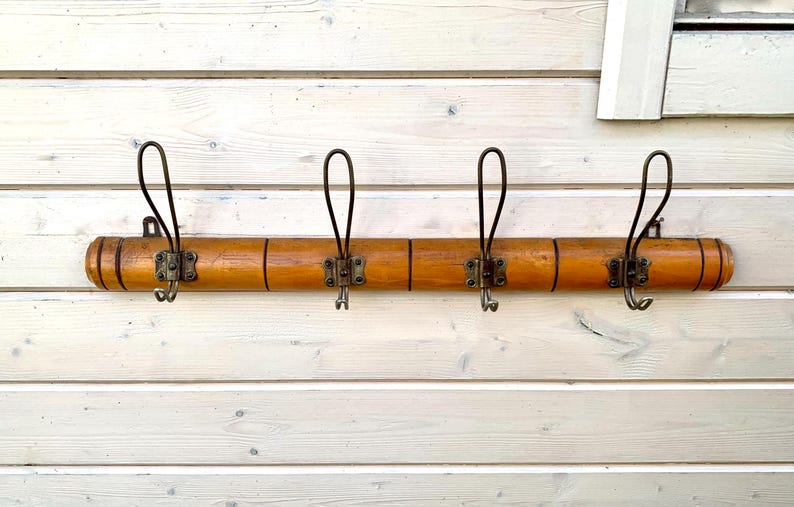 May include: A wooden coat rack with four hooks. The rack is made of light brown wood and has a natural finish. The hooks are made of metal and are black.