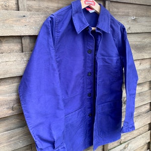 Pode incluir: Uma jaqueta de trabalho azul com fecho de bot&otilde;es e v&aacute;rios bolsos. A jaqueta est&aacute; pendurada em um cabide de madeira com a palavra "MASCOTTE" impressa nele.