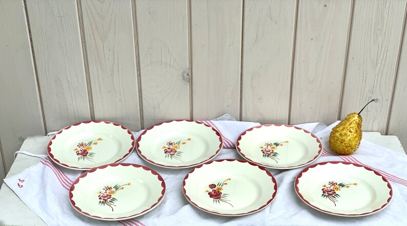 May include: Six white ceramic plates with a scalloped edge and a red trim. Each plate features a floral design with pink, yellow, and green flowers.