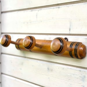 May include: Wooden coat rack with three pegs. The rack is made of light-coloured wood with dark brown stripes and accents. The pegs are round with a striped design. The rack is mounted on a white wooden wall.