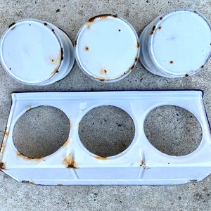 May include: Three white enamelware canisters with blue trim and rust spots, sitting on a white enamelware rack with three round openings.