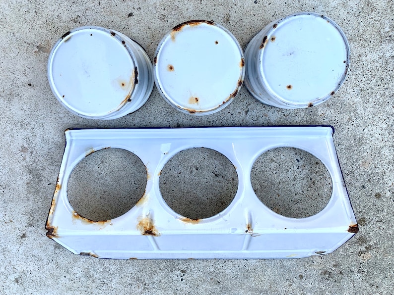 May include: Three white enamelware canisters with blue trim and rust spots, sitting on a white enamelware rack with three round openings.