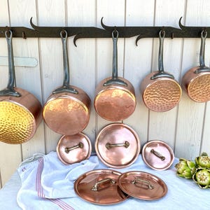 May include: A set of copper pans with hammered bases and silver handles, suspended from a wrought iron rack. Matching lids are also shown. The cookware is arranged on a white surface with a red striped towel and artichokes.