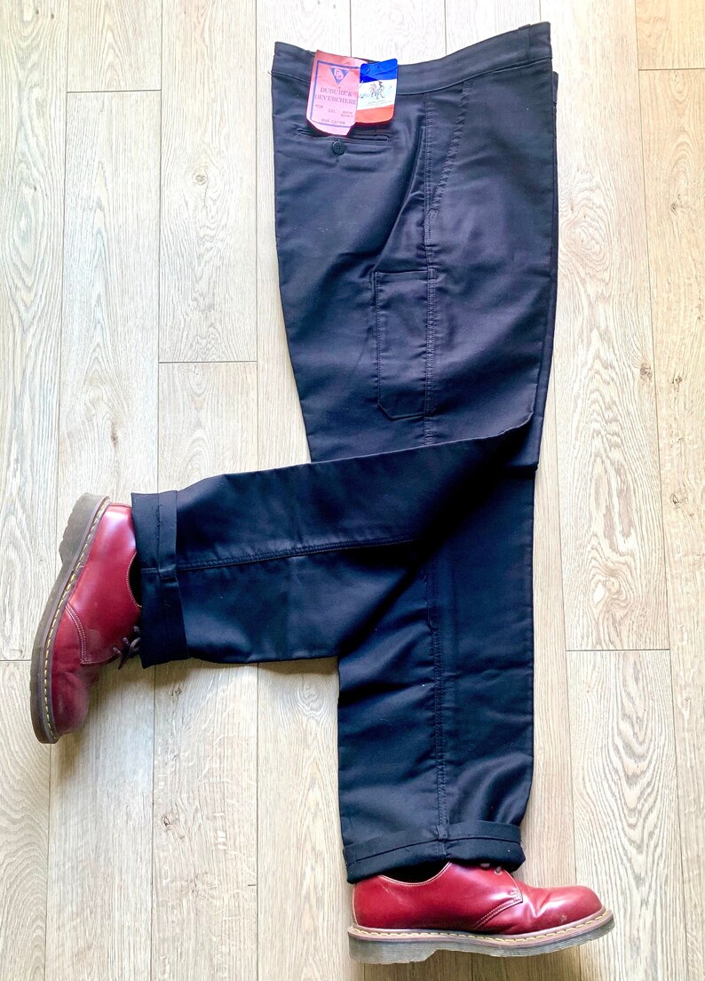 May include: A pair of dark blue cargo pants with a single large pocket on the right leg. The pants are folded and laid on a wooden floor with a pair of red leather shoes on either end.