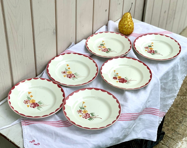 May include: Set of six white ceramic plates with a scalloped edge and a red rim. Each plate features a floral design with red, yellow, and green flowers.