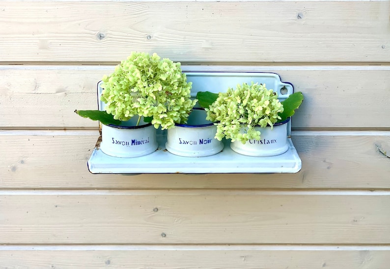 May include: Three white enamelware containers with blue trim, labeled "Savon Mineral", "Savon Noir", and "Cristaux", are mounted on a white shelf. Each container is filled with green hydrangea flowers.