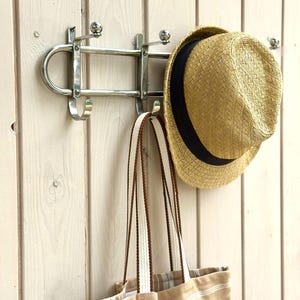Puede incluir: Un perchero de pared de metal plateado con varios ganchos. Un sombrero fedora de paja trenzada color canela con una cinta negra cuelga de un gancho, y un bolso de tela a rayas beige y blancas con asas trenzadas marrones cuelga de otro.