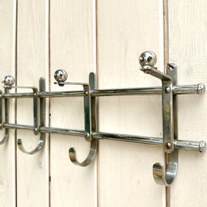 Puede incluir: Un perchero de pared fabricado en metal cromado pulido, con dos barras horizontales y cuatro soportes verticales. Cada soporte incluye un gancho curvado en la parte inferior y un remate esf&eacute;rico en forma de bola en la parte superior.