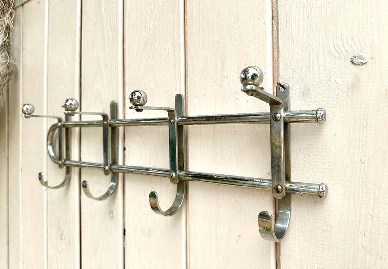 Puede incluir: Un perchero de pared fabricado en metal cromado pulido, con dos barras horizontales y cuatro soportes verticales. Cada soporte incluye un gancho curvado en la parte inferior y un remate esf&eacute;rico en forma de bola en la parte superior.