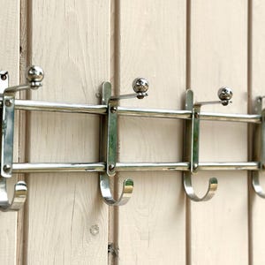 Puede incluir: Un perchero de pared hecho de metal plateado pulido, con un marco horizontal de doble barra, cuatro ganchos orientados hacia abajo y cuatro ganchos orientados hacia arriba rematados con peque&ntilde;as esferas para colgar prendas o accesorios.