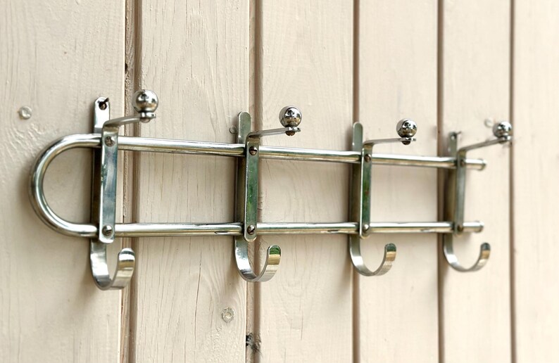Puede incluir: Un perchero de pared hecho de metal plateado pulido, con un marco horizontal de doble barra, cuatro ganchos orientados hacia abajo y cuatro ganchos orientados hacia arriba rematados con peque&ntilde;as esferas para colgar prendas o accesorios.