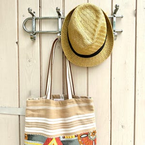 Puede incluir: Un sombrero fedora de paja con una banda negra y un bolso de tela rectangular con rayas horizontales beige y blancas en la parte superior y un patr&oacute;n geom&eacute;trico colorido en rojo, amarillo, naranja y gris en la parte inferior.
