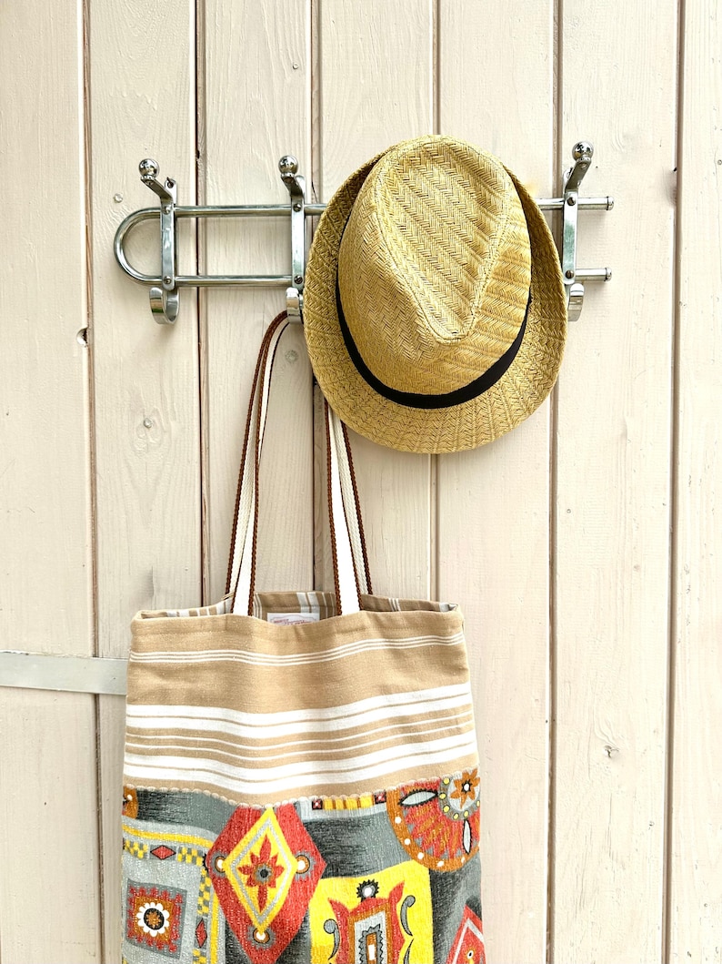 Puede incluir: Un sombrero fedora de paja con una banda negra y un bolso de tela rectangular con rayas horizontales beige y blancas en la parte superior y un patr&oacute;n geom&eacute;trico colorido en rojo, amarillo, naranja y gris en la parte inferior.