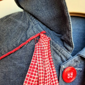 Puede incluir: Una chaqueta vaquera azul con ribete a cuadros rojo y blanco y un bot&oacute;n rojo.