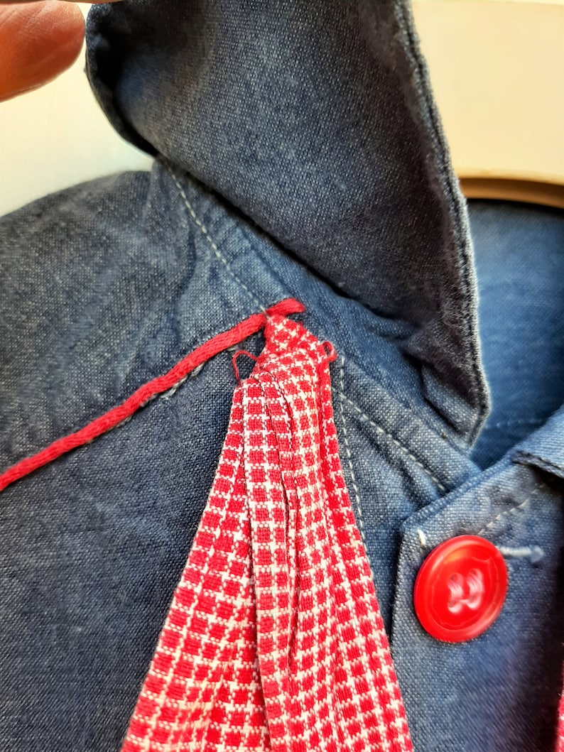 Puede incluir: Una chaqueta vaquera azul con ribete a cuadros rojo y blanco y un bot&oacute;n rojo.