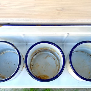 May include: White enamel wall mounted shelf with three blue rimmed round containers. The shelf has a blue trim and is slightly rusted.