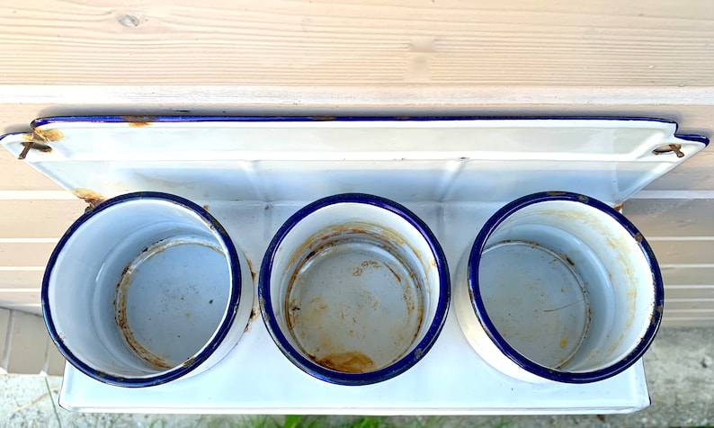 May include: White enamel wall mounted shelf with three blue rimmed round containers. The shelf has a blue trim and is slightly rusted.