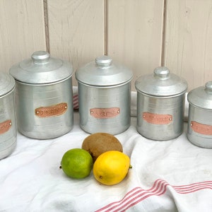 May include: Set of six silver metal canisters with copper labels. The labels read "Sugar", "Flour", "Rice", "Coffee", "Tea", and "Mince". The canisters are arranged in a row on a white and red striped tablecloth.
