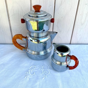 May include: A vintage chrome coffee pot set with bakelite handles and accents. The set includes a coffee pot, a creamer, and a lid. The coffee pot has a spout and a handle. The creamer has a handle. The set is displayed on a white tablecloth.