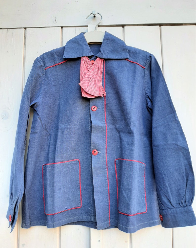 Puede incluir: Una camisa de algod&oacute;n azul con costuras rojas y una corbata de lazo a cuadros rojos y blancos. La camisa tiene dos bolsillos en la parte delantera y mangas largas.