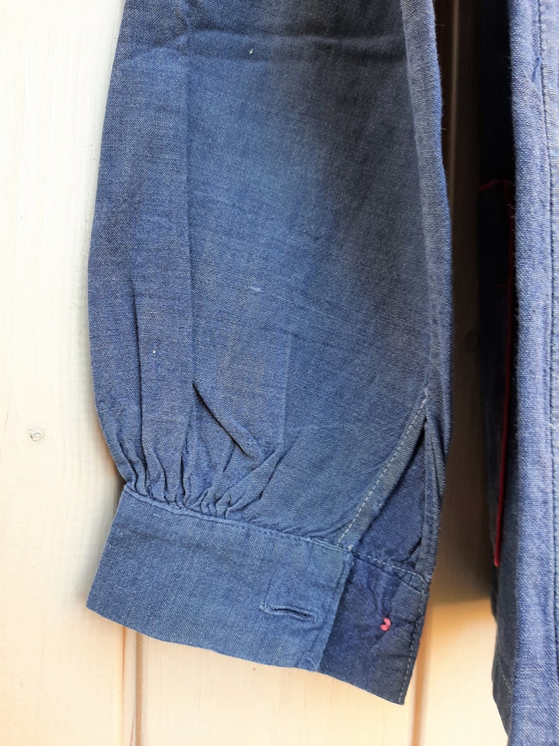 Puede incluir: Primer plano de una manga de camisa vaquera azul oscuro con un pu&ntilde;o fruncido y un cierre de bot&oacute;n. El pu&ntilde;o tiene un detalle de hilo rojo.