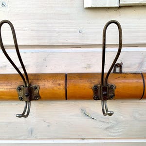 May include: A wooden coat rack with two metal hooks. The rack is made of light brown wood and has a natural finish. The hooks are made of metal and have a dark brown finish.