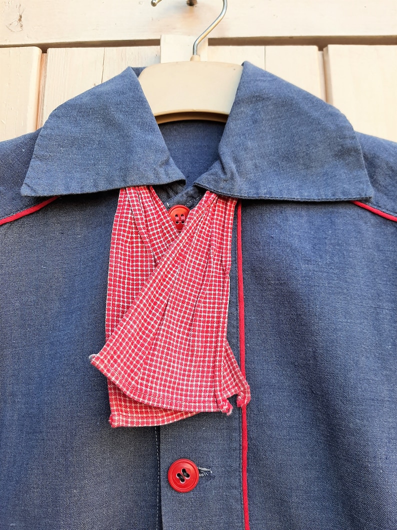 Puede incluir: Una camisa azul marino con una corbata de lazo a cuadros rojos y blancos. La camisa tiene ribetes rojos alrededor del cuello y en la parte delantera.