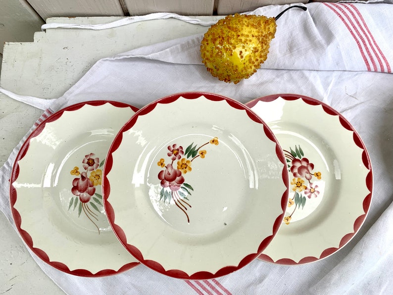 May include: Three white ceramic plates with red scalloped edges and floral designs in shades of red, yellow, and green.