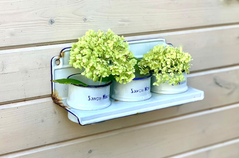 May include: White enamel shelf with three small white enamel cups with blue trim. Each cup holds a bunch of light green flowers. The cups have the text "Savon!" printed on them.