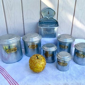 Peut inclure: Ensemble de sept boîtes en métal vintage avec couvercles. Les boîtes sont argentées et portent des étiquettes dorées avec les mots "Farine", "Sucre", "Café", "Thé", "Poivre", "Épices" et "Biscuits".
