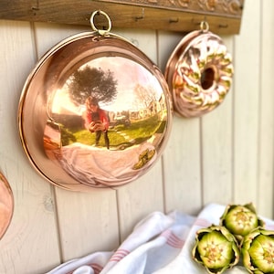 Pode incluir: Utensílios de cozinha de cobre pendurados em um suporte de madeira. A imagem apresenta uma panela grande e redonda de cobre com uma superfície reflexiva, uma forma de bolo e um suporte de madeira decorativo. Um pano branco e vermelho listrado e alcachofras estão em primeiro plano.