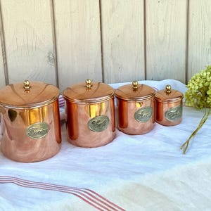 May include: Four copper kitchen canisters with lids, labelled "Farine", "Café", "Sucre", and "Sel". The canisters are on a white cloth with red stripes. A small bouquet of dried flowers is to the right.