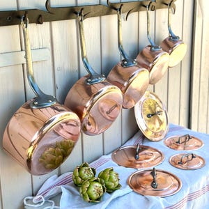 May include: A collection of five polished copper saucepans with long metal handles hanging from a black iron wall rack. Three matching copper lids with metal handles are placed on a white cloth, alongside three fresh green artichokes.