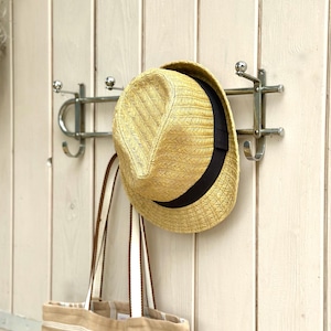 Puede incluir: Un sombrero fedora de paja con una banda negra y un bolso de tela a rayas beige y blanco colgados de un perchero de pared de metal plateado con varios ganchos. El perchero presenta un dise&ntilde;o de barra horizontal con remates redondeados.