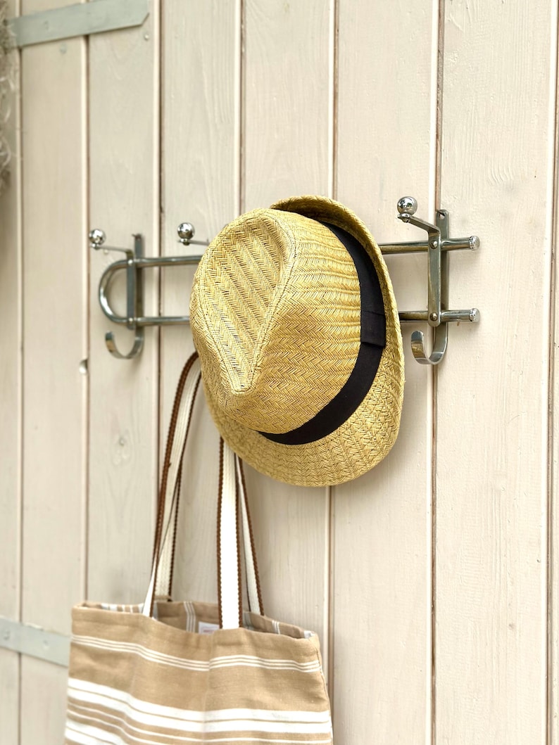 Puede incluir: Un sombrero fedora de paja con una banda negra y un bolso de tela a rayas beige y blanco colgados de un perchero de pared de metal plateado con varios ganchos. El perchero presenta un dise&ntilde;o de barra horizontal con remates redondeados.
