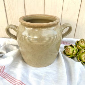 Op de afbeelding: Een beige antieke keramische pot met twee handvatten en horizontale groeven. De pot heeft een lichtbruine rand en de letter "S" is zichtbaar. Drie artisjokken liggen op een witte doek met rode strepen.