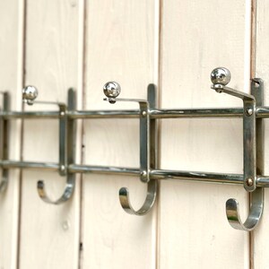Puede incluir: Un perchero de pared hecho de metal cromado pulido, con cuatro ganchos curvos para colgar objetos. El perchero presenta un dise&ntilde;o de doble barra horizontal con peque&ntilde;os remates esf&eacute;ricos en la parte superior de cada soporte.