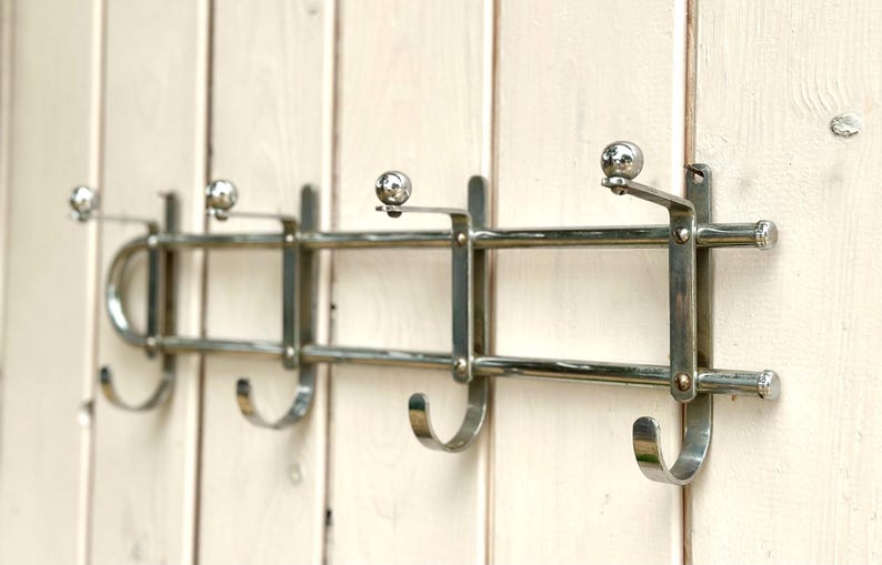 Puede incluir: Un perchero de pared hecho de metal cromado pulido, con cuatro ganchos curvos para colgar objetos. El perchero presenta un dise&ntilde;o de doble barra horizontal con peque&ntilde;os remates esf&eacute;ricos en la parte superior de cada soporte.