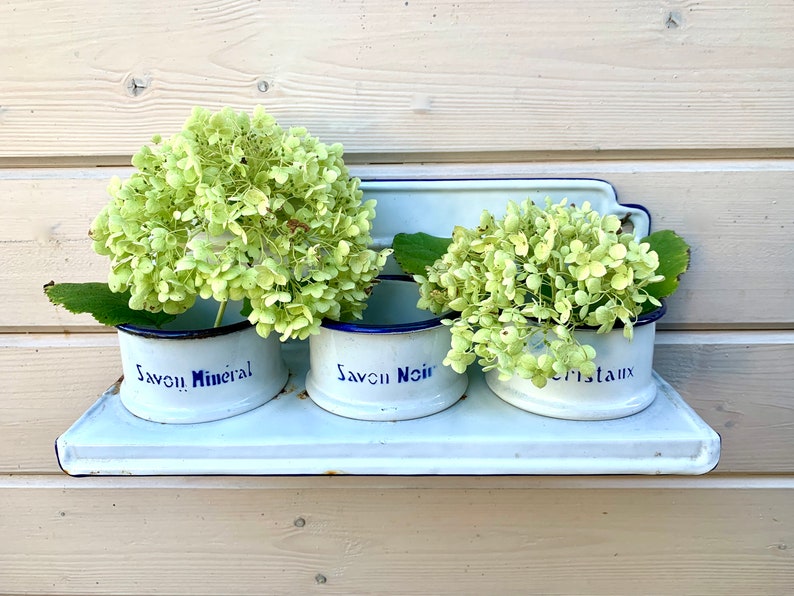 May include: Three white enamelware containers with blue trim, labeled "Savon Mineral", "Savon Noir", and "Cristaux", are mounted on a white shelf. Each container is filled with a bouquet of light green hydrangeas.