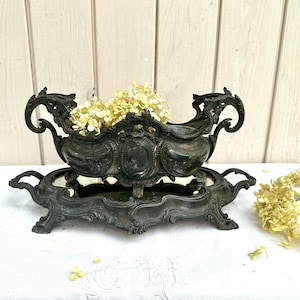 May include: A dark metallic centrepiece bowl with ornate scrollwork handles and a central oval medallion, resting on a matching rectangular mirrored tray with decorative feet. The bowl contains dried cream-coloured hydrangea flower clusters.