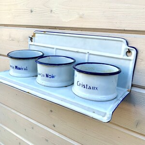 May include: White enamel shelf with three white enamel containers with blue rims and blue lettering. The containers are labeled "Sel Mineral", "Savon Noir", and "Cristaux".