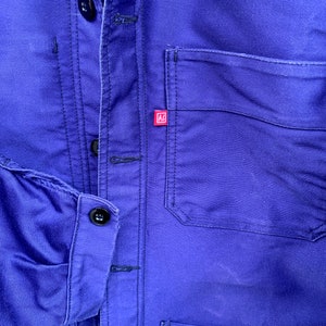 Pode incluir: Uma jaqueta de trabalho azul com um remendo vermelho no peito. A jaqueta tem fecho de bot&otilde;es e v&aacute;rios bolsos.