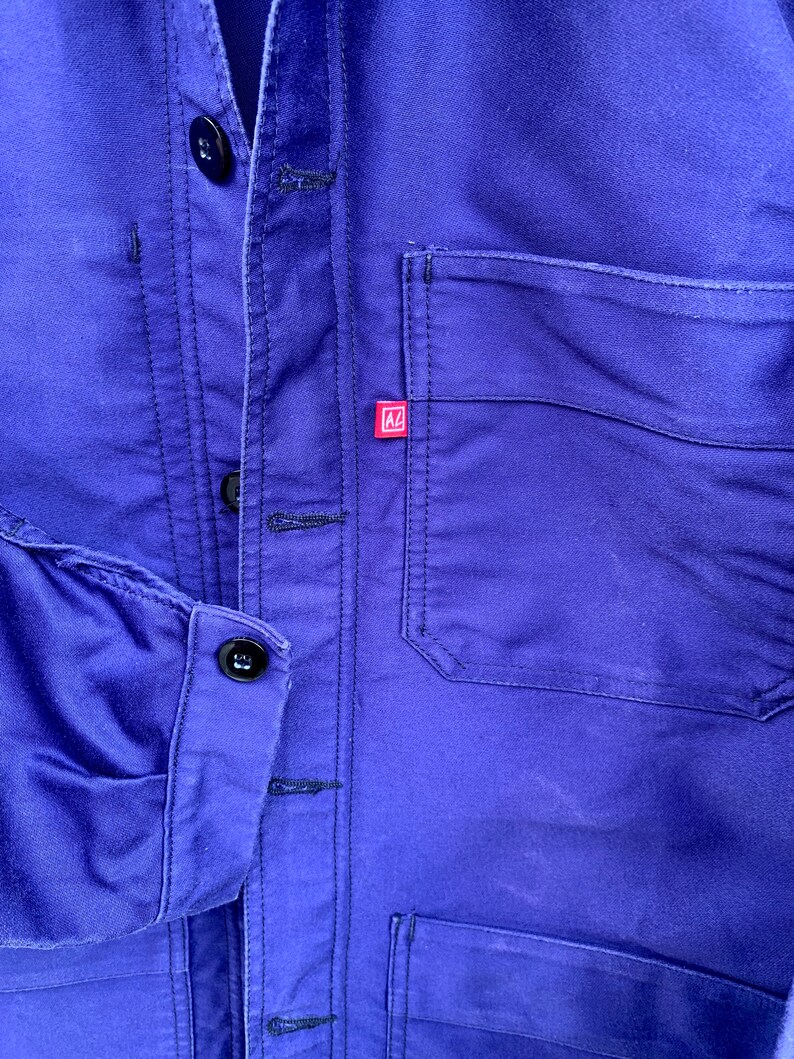 Pode incluir: Uma jaqueta de trabalho azul com um remendo vermelho no peito. A jaqueta tem fecho de bot&otilde;es e v&aacute;rios bolsos.