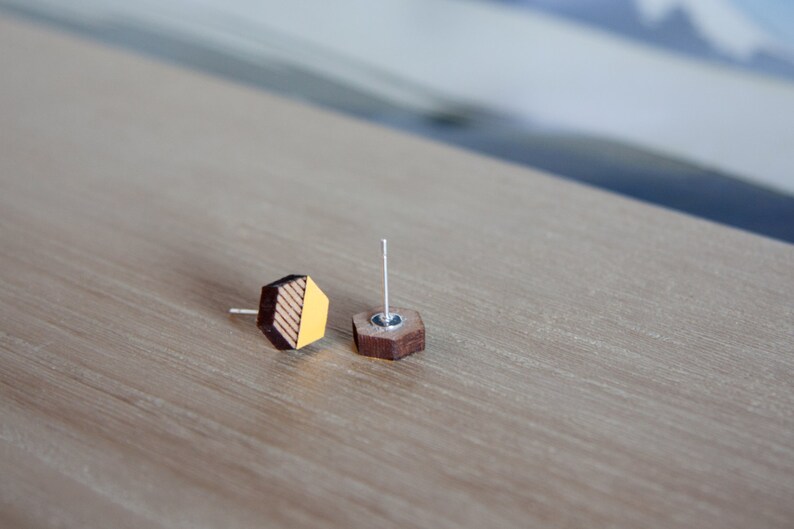 May include: A pair of hexagonal wooden earrings with a yellow and brown striped design. The earrings are on a light brown wooden surface.