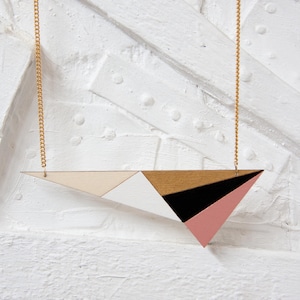 Puede incluir: Un collar con un colgante geométrico y una cadena dorada. El colgante está hecho de madera y pintado en tonos de rosa, blanco, negro y dorado.