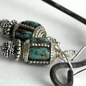 May include: Close-up of a pair of silver earrings with turquoise-colored stone beads. The earrings have intricate silver detailing and a decorative design. The earrings are on a white surface.