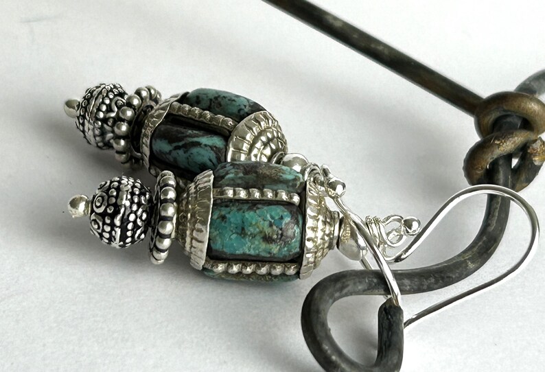 May include: Close-up of a pair of silver earrings with turquoise-colored stone beads. The earrings have intricate silver detailing and a decorative design. The earrings are on a white surface.