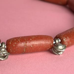 Pode incluir: Close-up de um colar com contas cil&iacute;ndricas castanho-avermelhadas com uma textura salpicada. As contas s&atilde;o conectadas por pequenos pingentes de metal prateado. O colar &eacute; exibido em um fundo rosa.