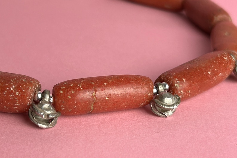 Pode incluir: Close-up de um colar com contas cil&iacute;ndricas castanho-avermelhadas com uma textura salpicada. As contas s&atilde;o conectadas por pequenos pingentes de metal prateado. O colar &eacute; exibido em um fundo rosa.